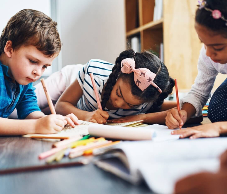 3 children, one boy and two girls are drawing in their notebooks. Art supplies are kept in front of them.