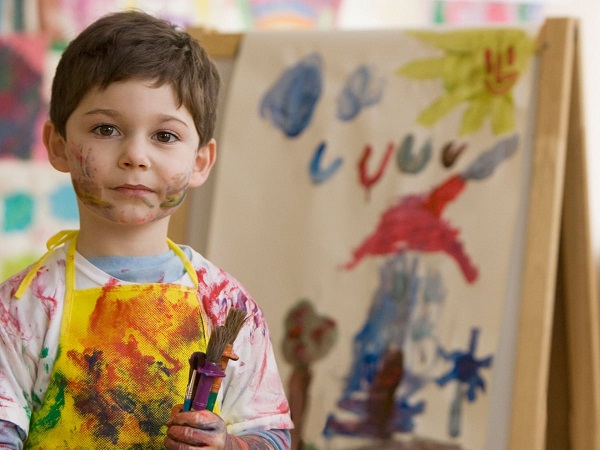 A boy is standing in front of a painting done by him. he has brushes in his hand and his dress and face are smeared with paint